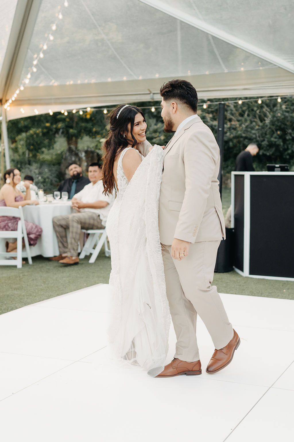 A couple dressed elegantly shares a dance on a white platform under a clear tent, with seated guests in the background at Kualoa ranch