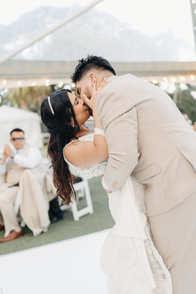 A couple in wedding attire embraces and kisses on a dance floor. Guests in the background sit at tables under a tent.