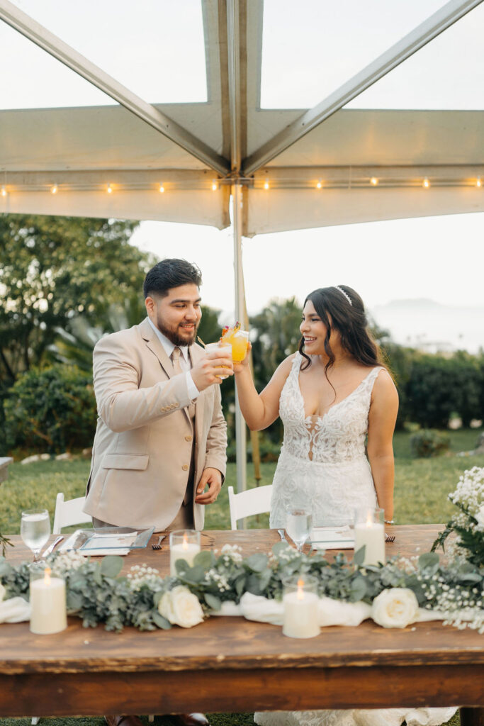 A couple in formal attire clinks their glasses under a tent at an outdoor event, with a decorated wooden table in the foreground.