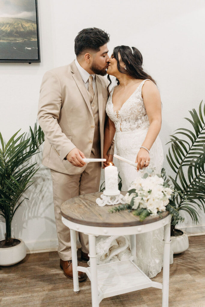 A bride and groom share a kiss while holding candles together over a small table with a floral centerpiece.