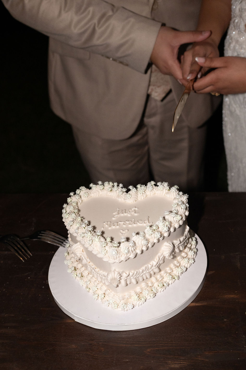 A heart-shaped cake with white frosting and decorative pearls on a table; a couple holding a knife stands in the background.