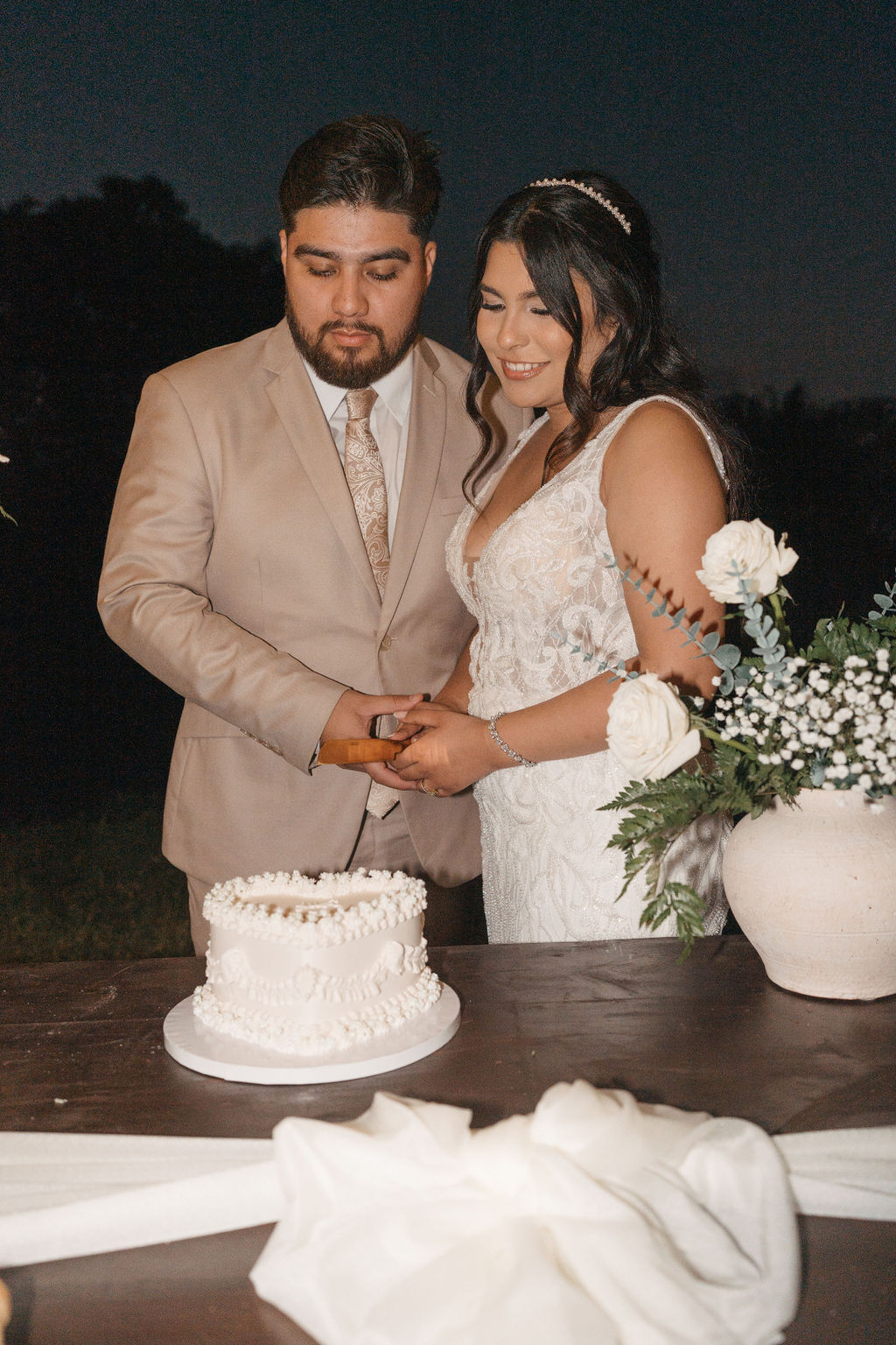 A heart-shaped cake with white frosting and decorative pearls on a table; a couple holding a knife stands in the background.