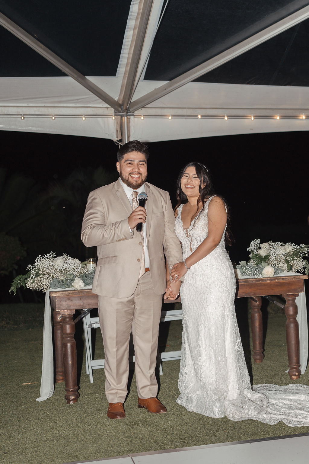 A couple stands together under a tent at night. The man holds a microphone, and both are smiling. They are in formal attire, with the woman in a white dress. A table with flowers is behind them.