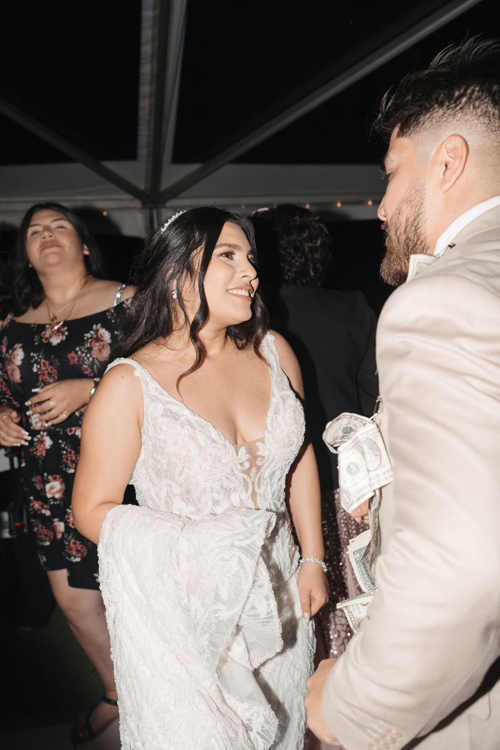 A bride in a white dress talks to a man in a light suit holding cash. Another person in a floral dress is in the background.