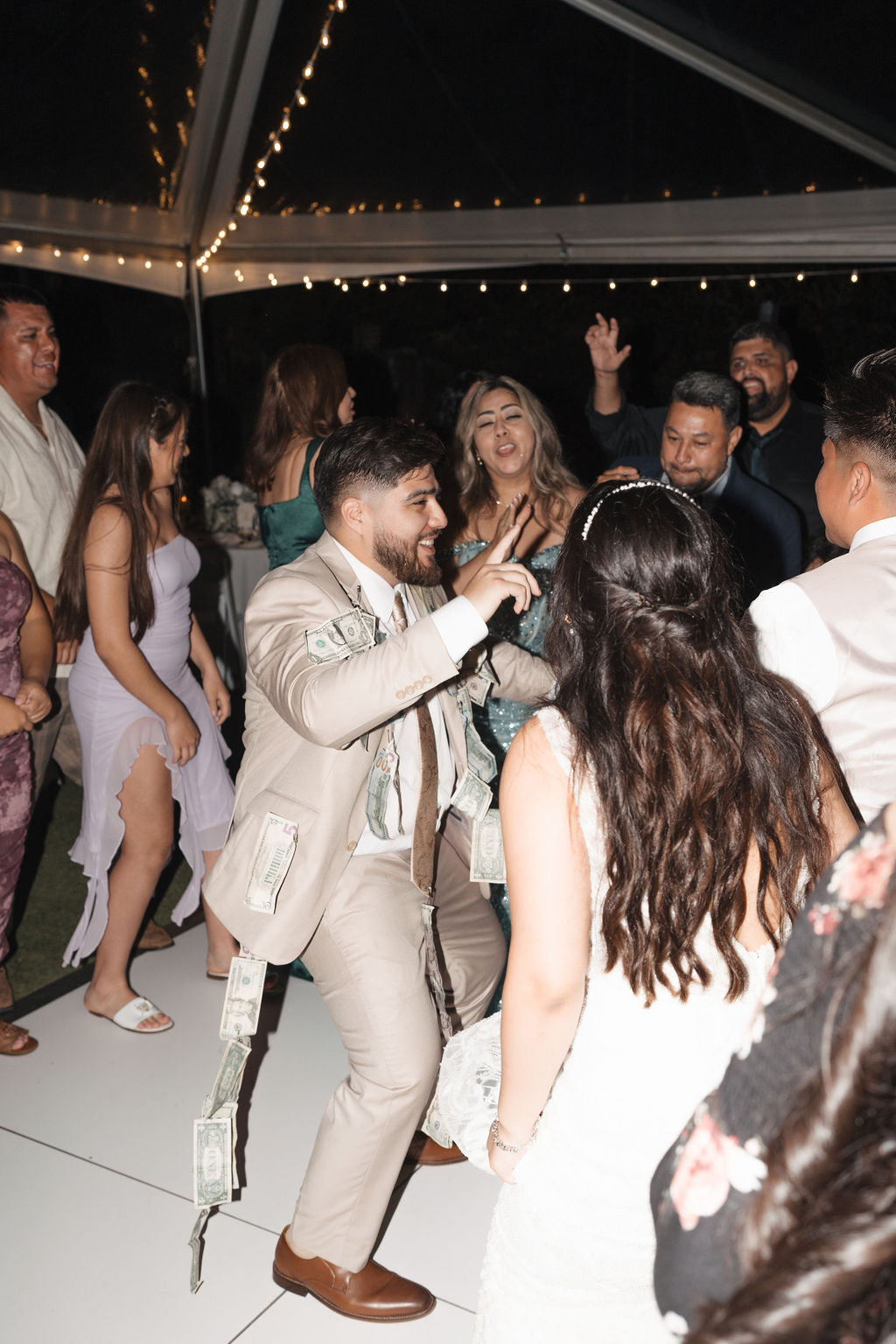 A group of people, some wearing formal attire, are dancing energetically under string lights. One person has paper bills attached to their suit.