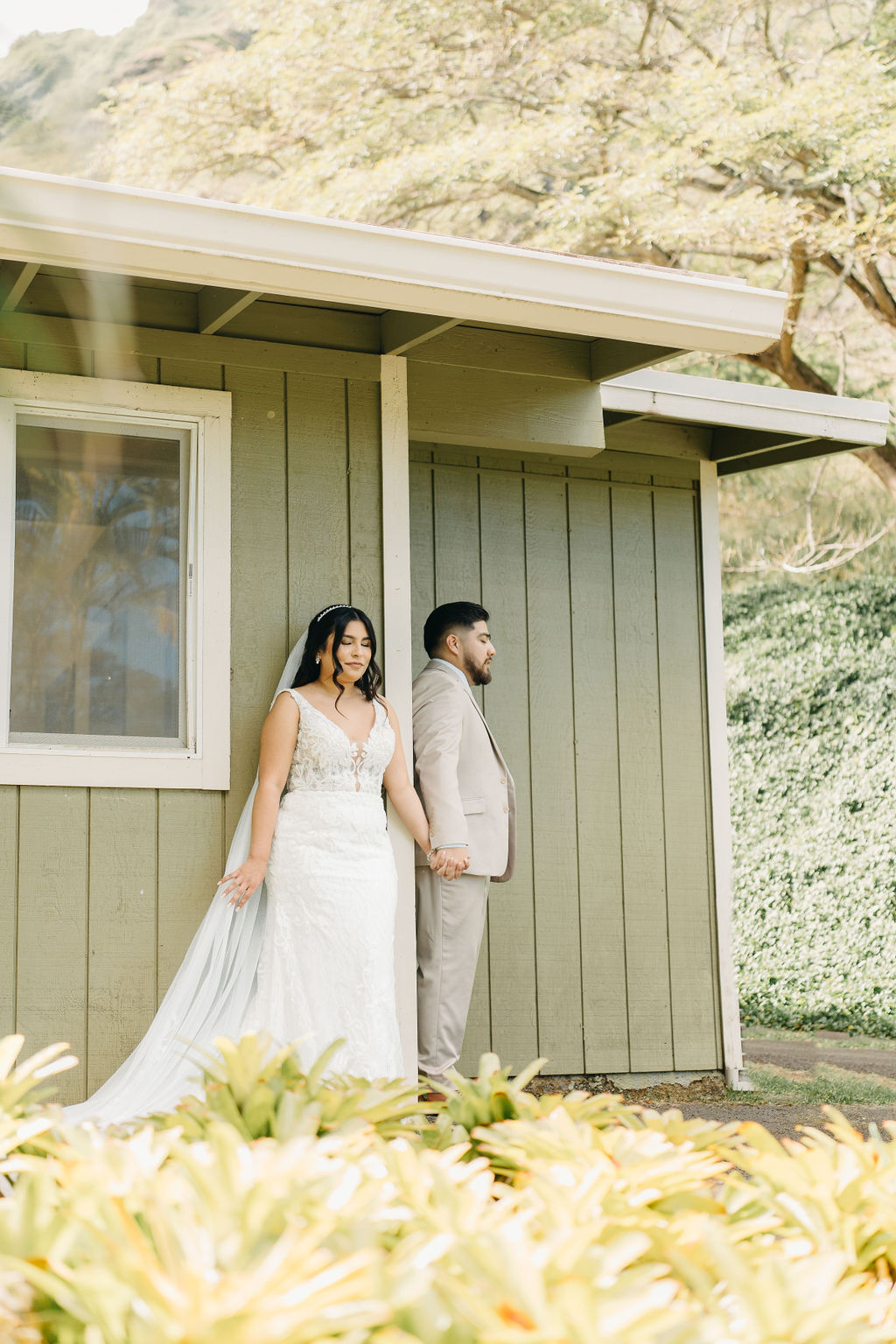 A bride and groom stand back-to-back and hold hands, facing away from each other, beside a green building with plants in the foreground.