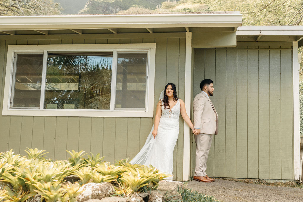 A bride and groom stand back-to-back and hold hands, facing away from each other, beside a green building with plants in the foreground.