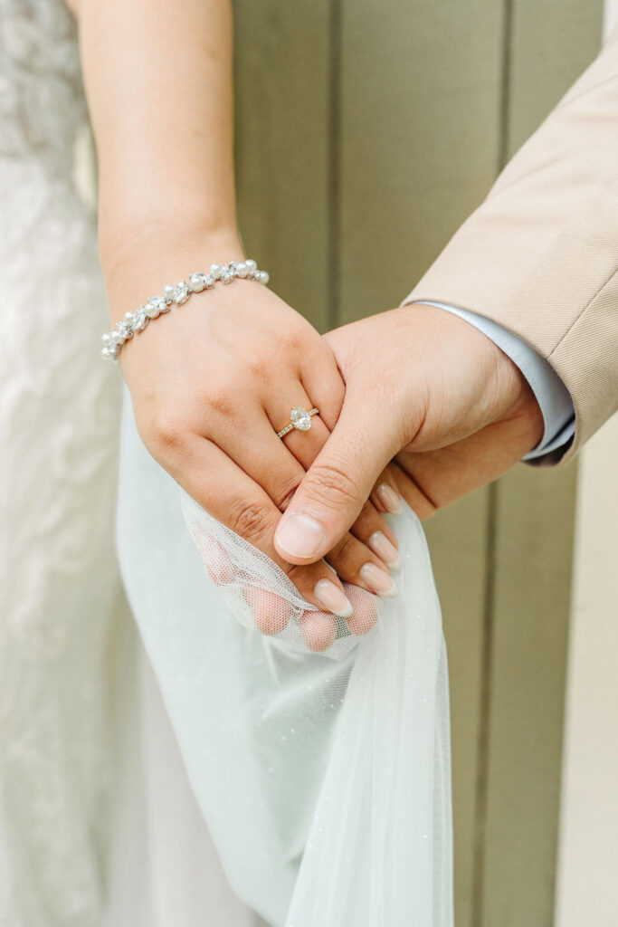 A close-up of two people holding hands, one wearing a pearl bracelet and wedding ring, the other in a beige suit, grasping light fabric.