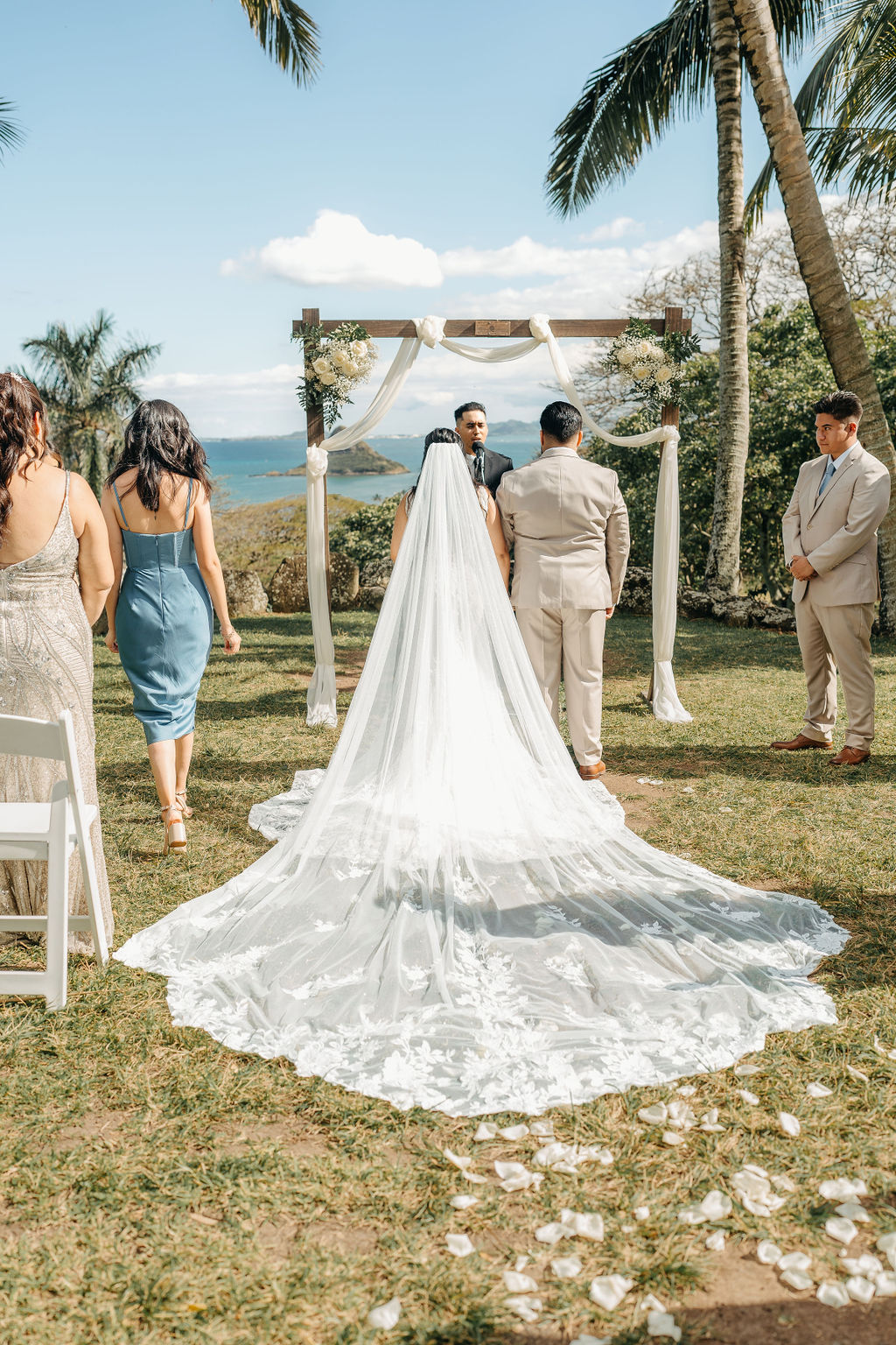 A bride walks down the aisle outdoors with a long veil, approaching the altar where the groom and officiant await. Guests stand nearby under palm trees at kualoa ranch