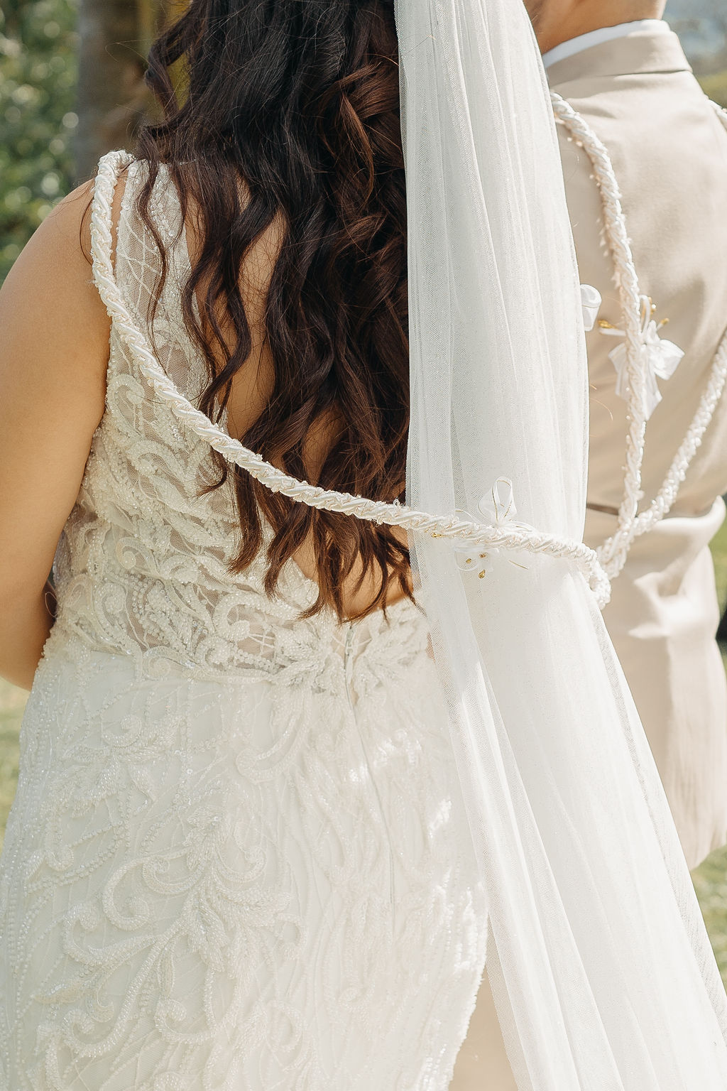 A bride walks down the aisle outdoors with a long veil, approaching the altar where the groom and officiant await. Guests stand nearby under palm trees at kualoa ranch