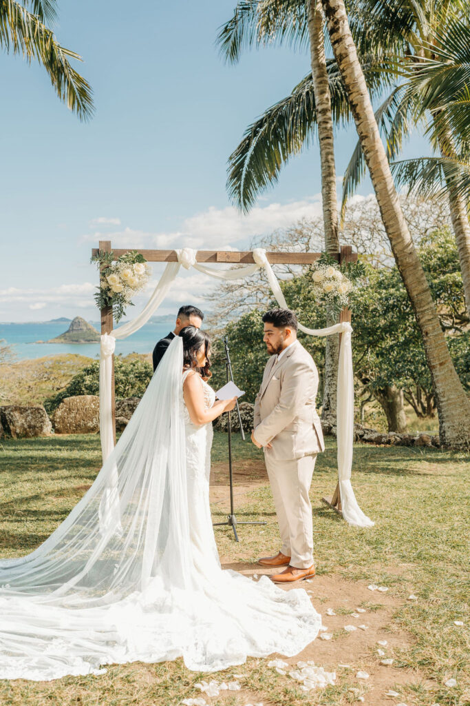 A couple stands beneath a decorated wedding arch outdoors, with palm trees and distant water visible. The bride holds a paper, possibly reading vows, while the groom listens attentively.