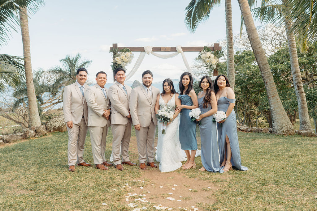 A wedding party poses outdoors on grass. The group includes the bride, groom, bridesmaids in blue dresses, and groomsmen in beige suits, with palm trees and a decorated arch in the background.