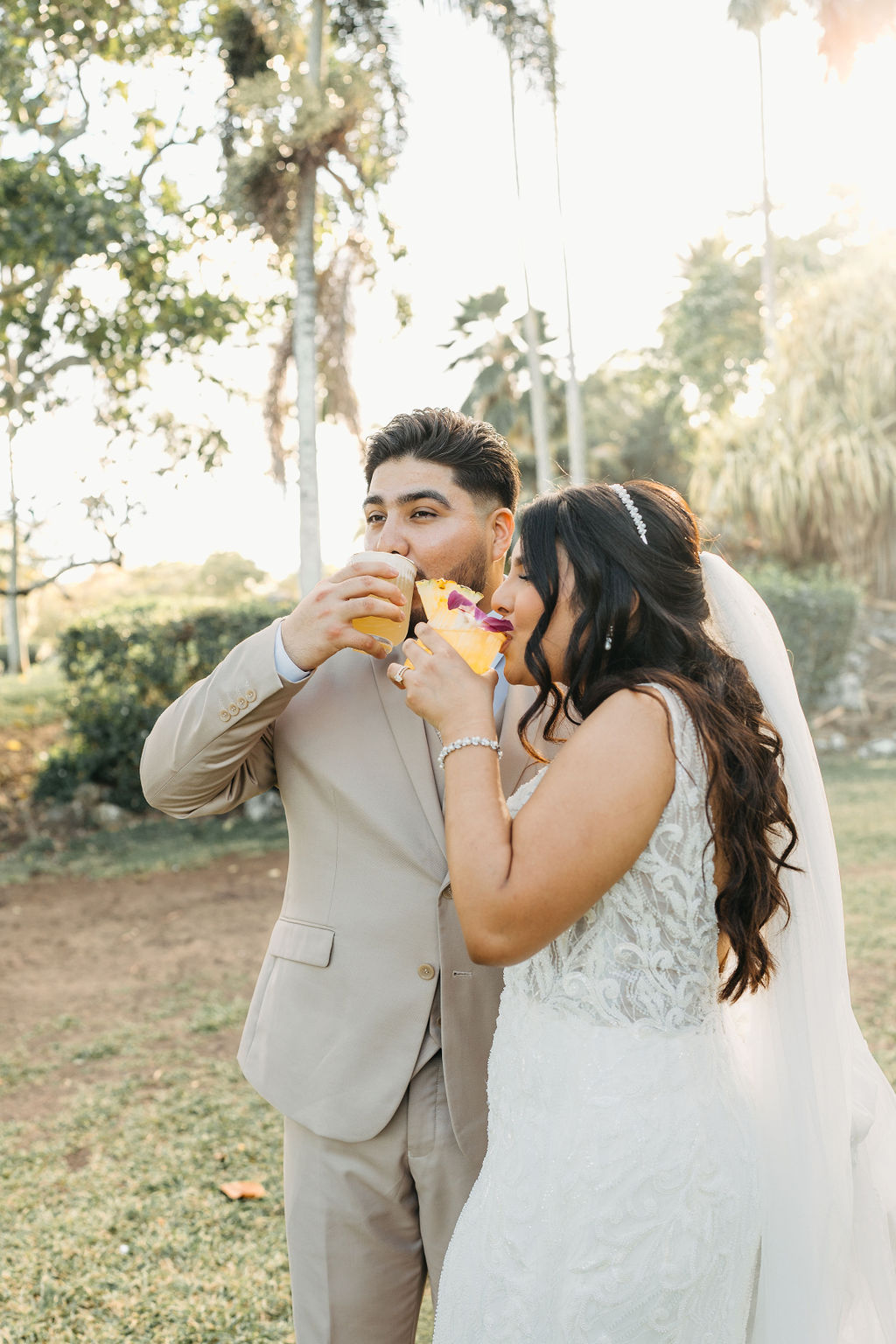 A bride and groom in wedding attire sip drinks outdoors, surrounded by trees and greenery.