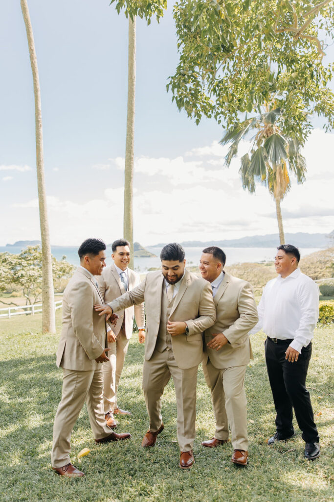 Five men in suits and dress shoes share a joyful moment outdoors on a grassy area with palm trees and a distant ocean view.