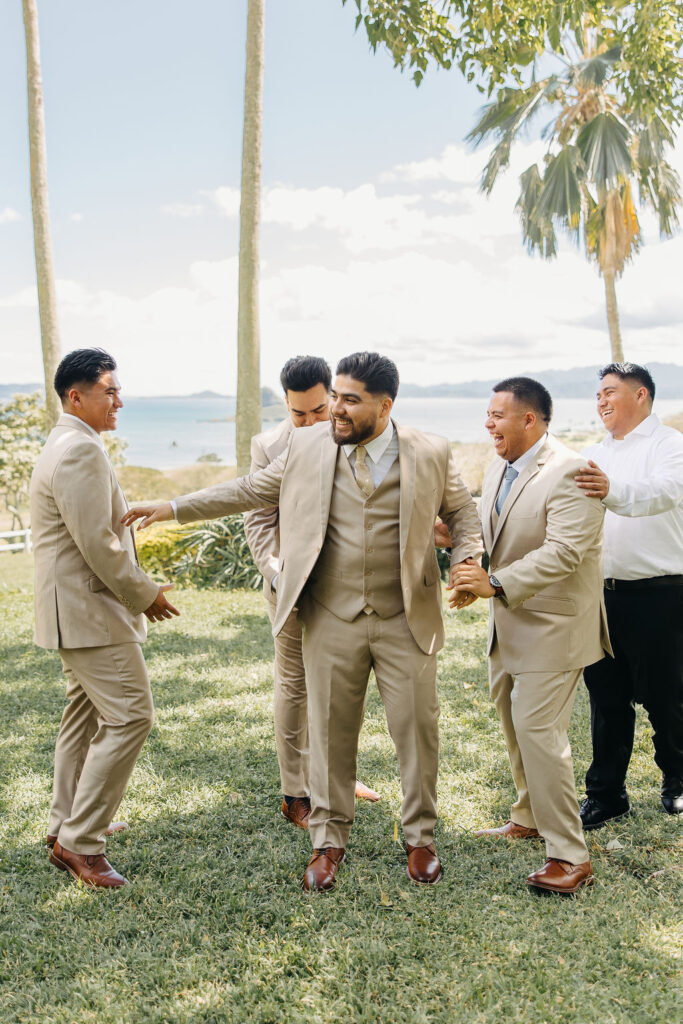 Five men in suits and dress shoes share a joyful moment outdoors on a grassy area with palm trees and a distant ocean view.