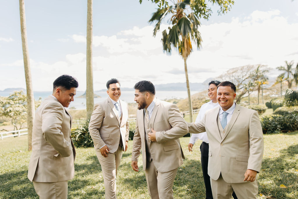 Five men in suits and dress shoes share a joyful moment outdoors on a grassy area with palm trees and a distant ocean view.