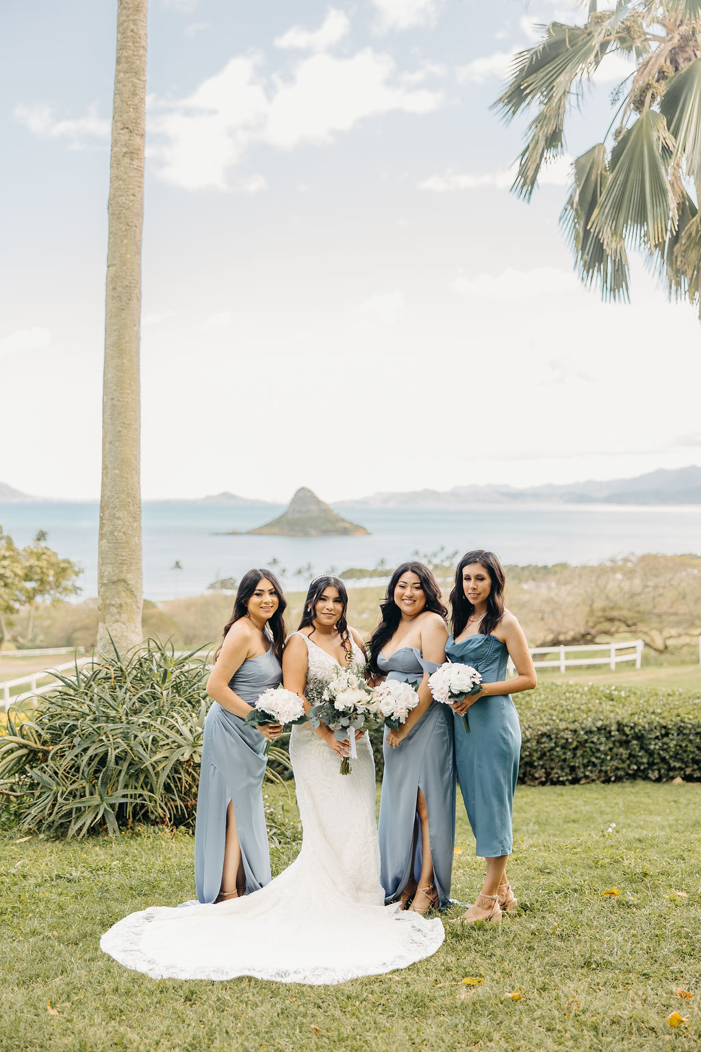 A wedding party poses outdoors on grass. The group includes the bride, groom, bridesmaids in blue dresses, and groomsmen in beige suits, with palm trees and a decorated arch in the background.