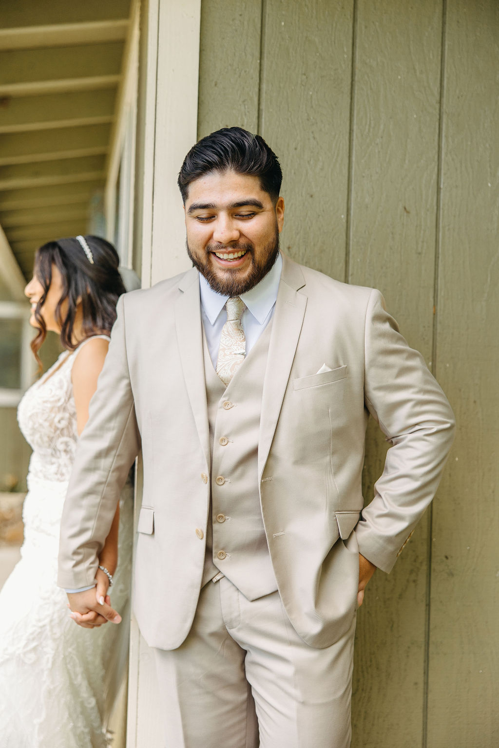 A bride and groom stand back-to-back and hold hands, facing away from each other, beside a green building with plants in the foreground.