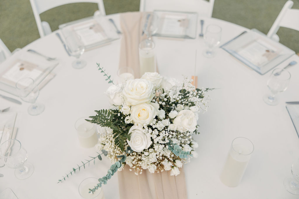 A round table is set with white chairs, glassware, and napkins. The centerpiece is an arrangement of white roses, baby's breath, and greenery on a beige table runner.