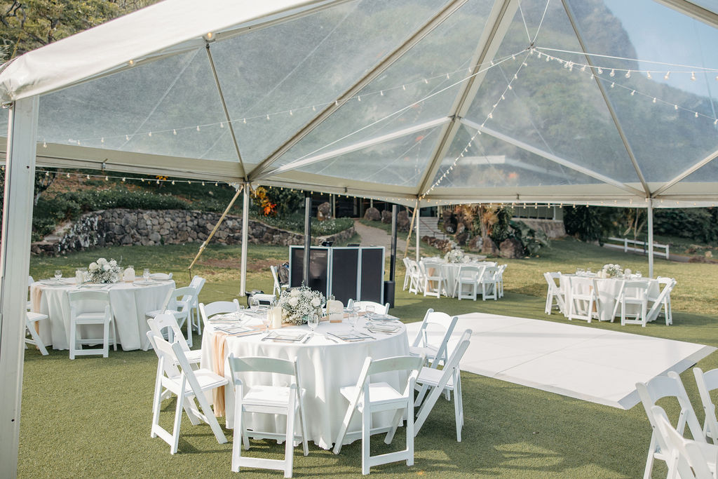 Outdoor event with a white tent set up on a grassy area surrounded by trees and palm plants under a clear sky at Kualoa ranch 