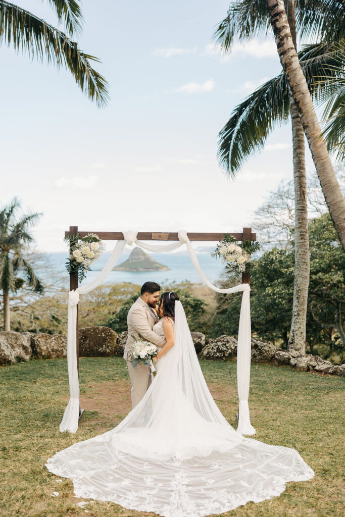 A couple stands beneath a decorated wedding arch outdoors, with palm trees and distant water visible. The bride holds a paper, possibly reading vows, while the groom listens attentively.