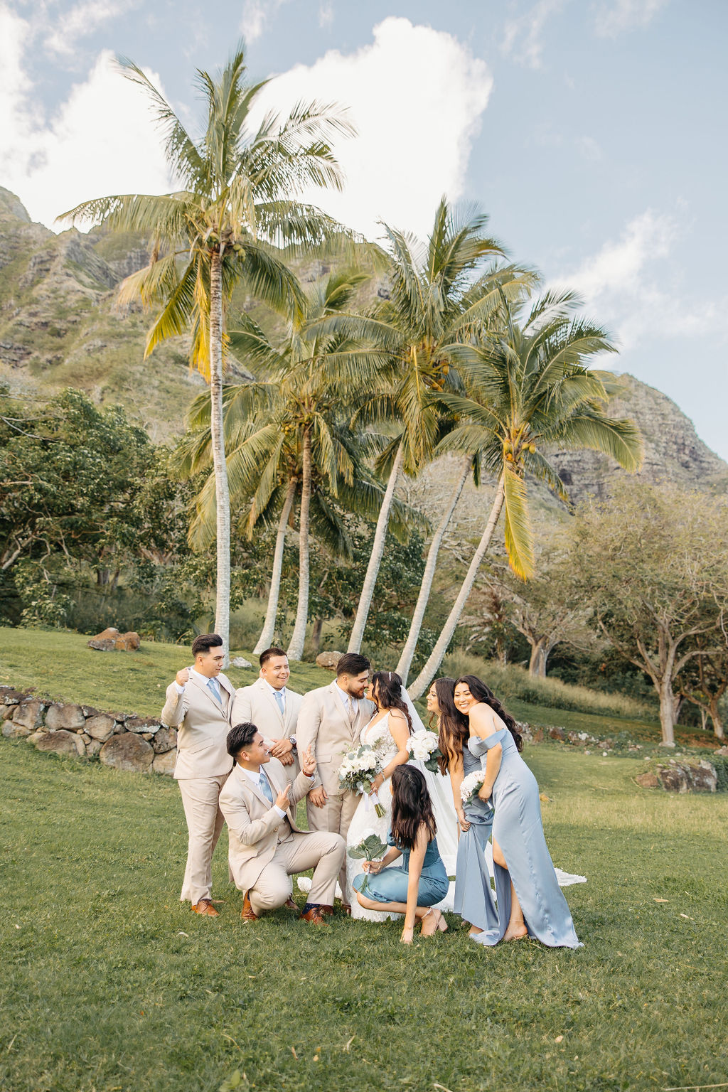 A wedding party poses outdoors on grass. The group includes the bride, groom, bridesmaids in blue dresses, and groomsmen in beige suits, with palm trees and a decorated arch in the background.