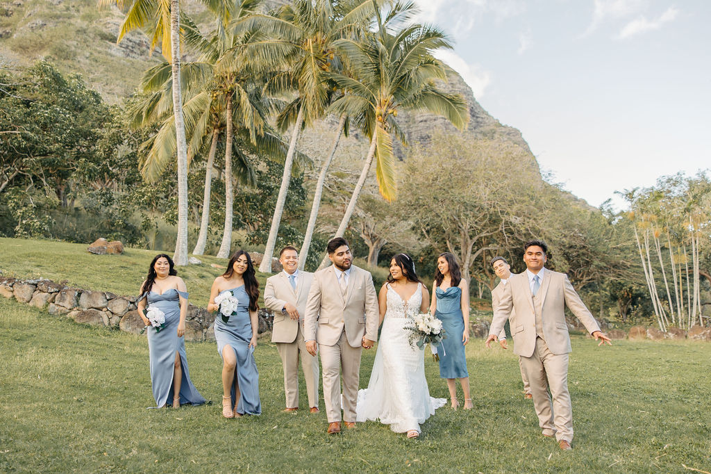 A wedding party poses outdoors on grass. The group includes the bride, groom, bridesmaids in blue dresses, and groomsmen in beige suits, with palm trees and a decorated arch in the background.