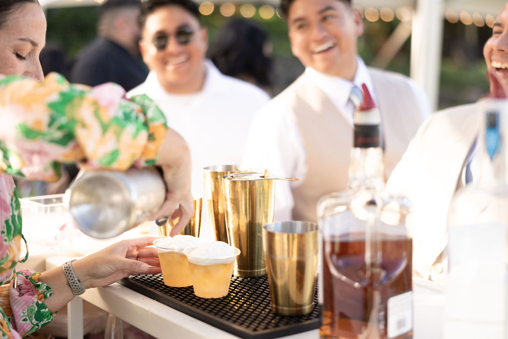 A person pours drinks into cups at a bar with golden shakers, while three people in the background are smiling.