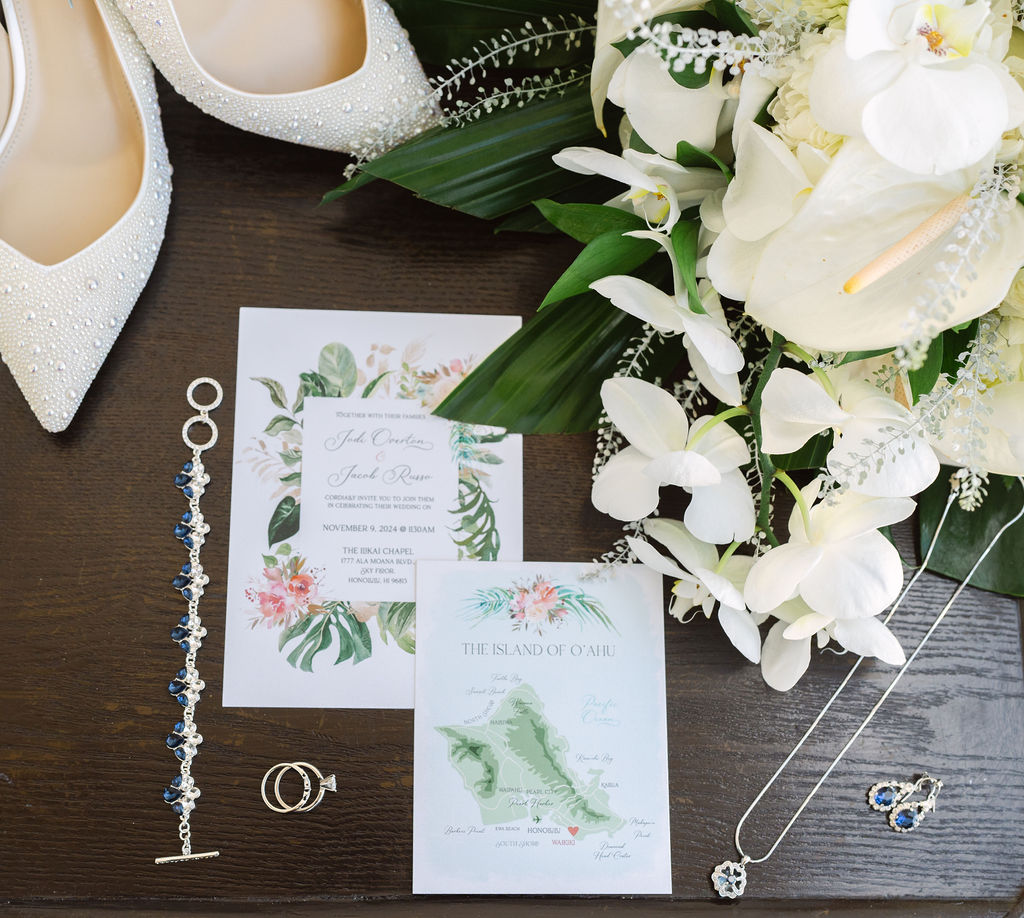 Wedding invitation with floral design on a table surrounded by white flowers, a pair of white shoes, a bracelet, rings, and a necklace.