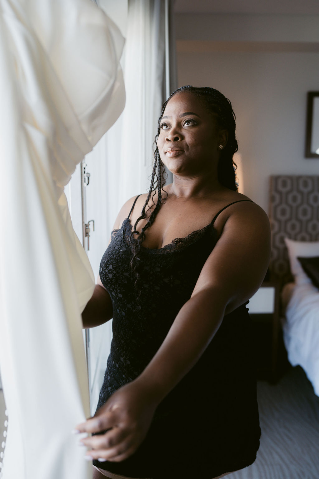 A woman in a black dress holds and looks at a white garment near a window in a bright room for a micro wedding in Oahu