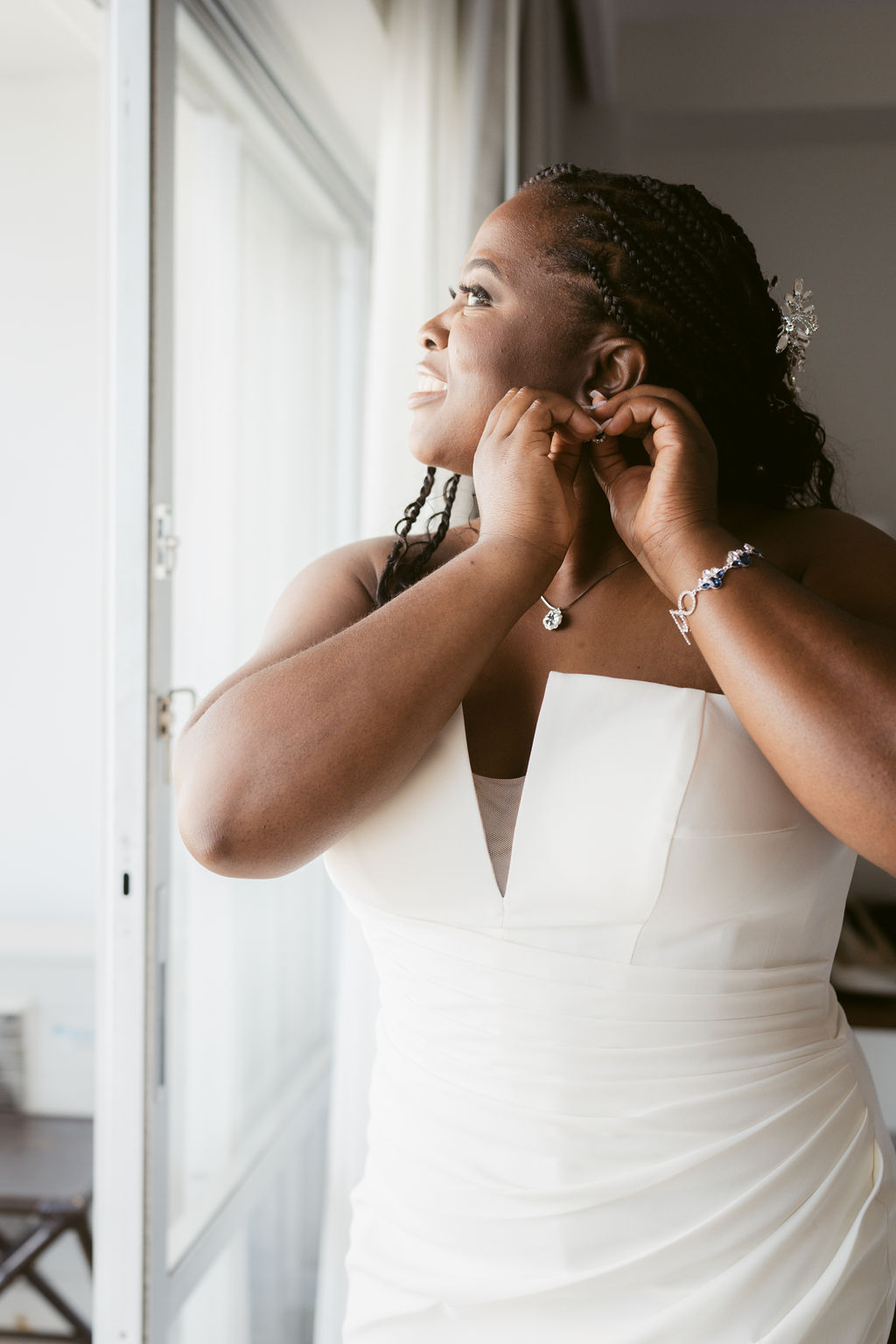 Bride in a white dress looks out a window while adjusting her earring.