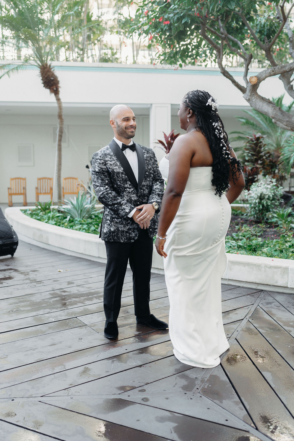 A couple stands facing each other outdoors. The man wears a patterned suit, and the woman is in a white dress. There are trees and plants in the background.