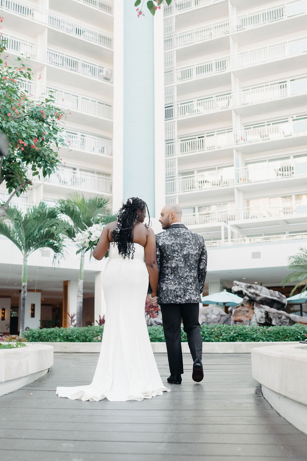 A couple stands facing each other outdoors. The man wears a patterned suit, and the woman is in a white dress. There are trees and plants in the background.