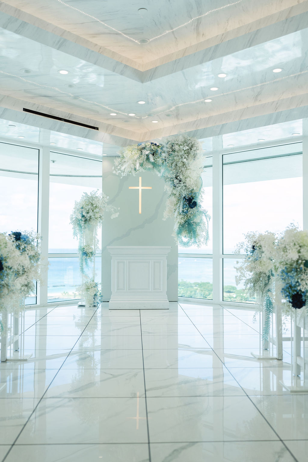 A bright, modern chapel with large windows, white marble flooring, a white altar with a cross, and floral arrangements in blue and white tones.