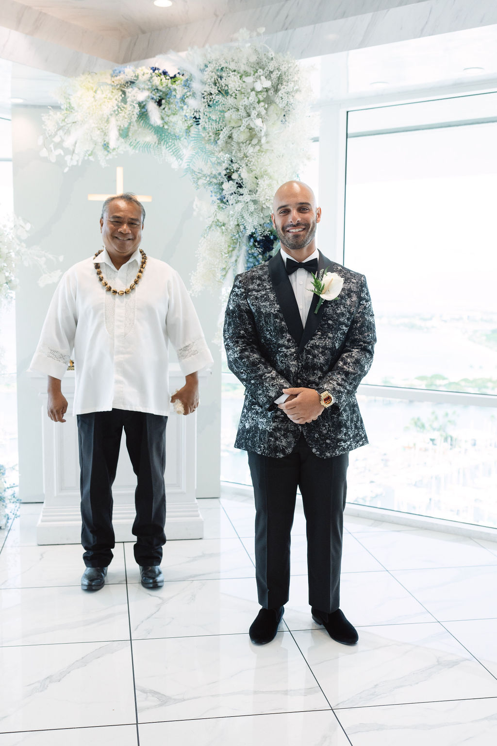 Two men stand indoors; one in a floral suit with a boutonniere, the other in a casual white shirt and lei. They're positioned on a glossy floor with a floral arrangement behind them.