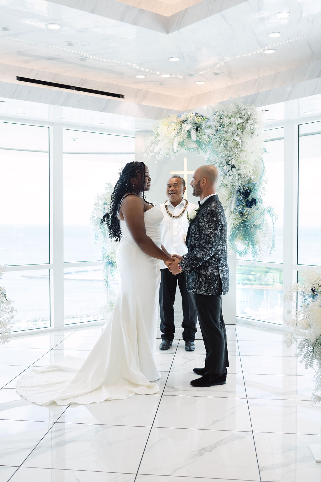 A couple holds hands and exchanges vows in a bright, modern room with large windows and floral decorations, while an officiant stands behind them.