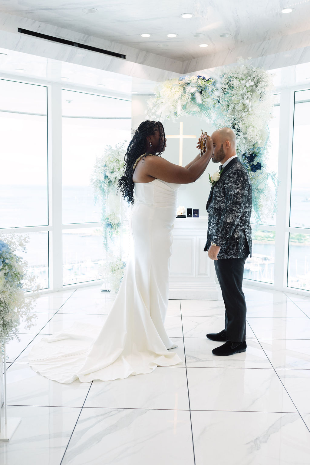 A couple holds hands and exchanges vows in a bright, modern room with large windows and floral decorations, while an officiant stands behind them for a micro wedding in oahu