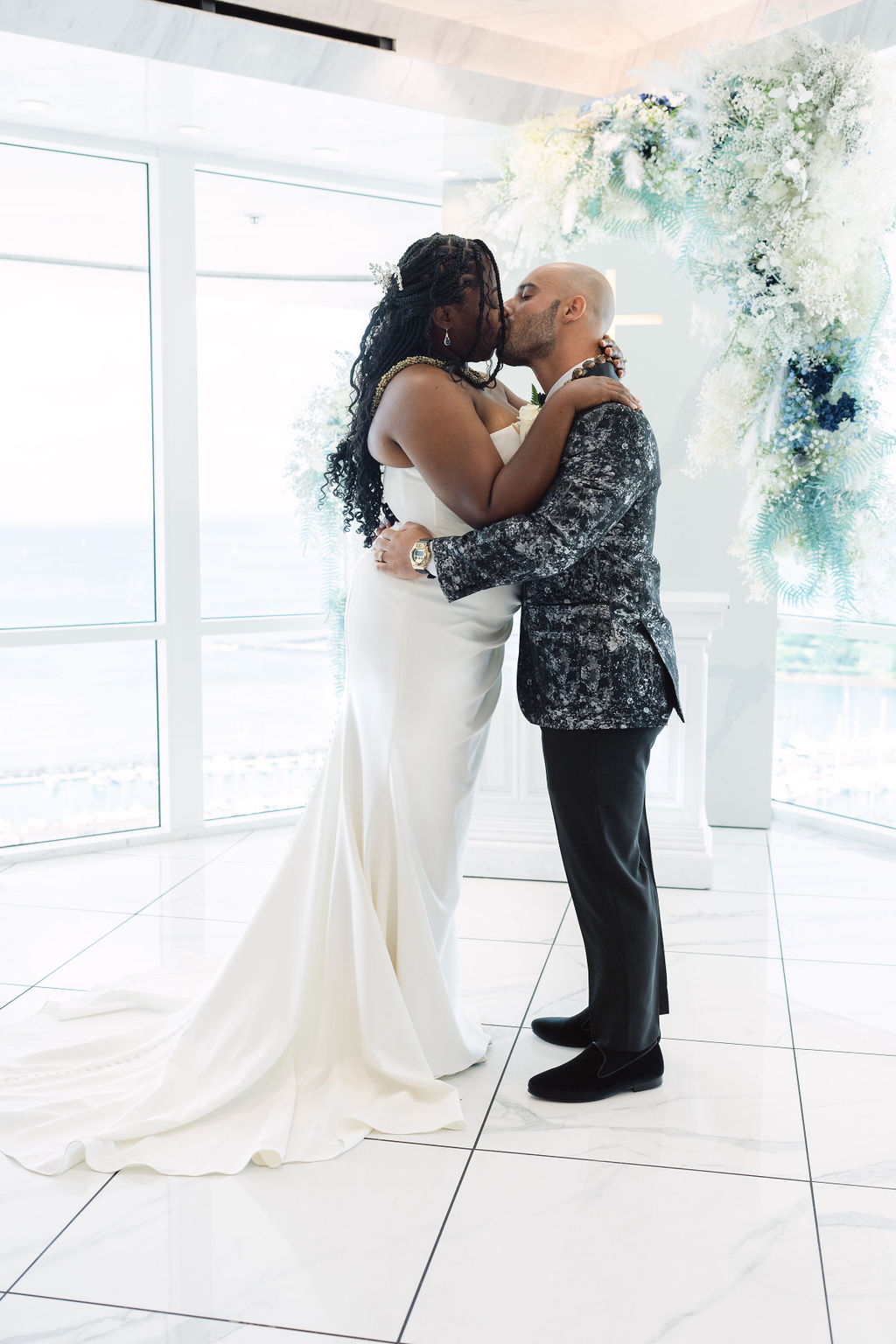 A couple holds hands and exchanges vows in a bright, modern room with large windows and floral decorations, while an officiant stands behind them for a micro wedding in oahu