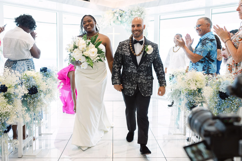 A couple holds hands and exchanges vows in a bright, modern room with large windows and floral decorations, while an officiant stands behind them for a micro wedding in oahu