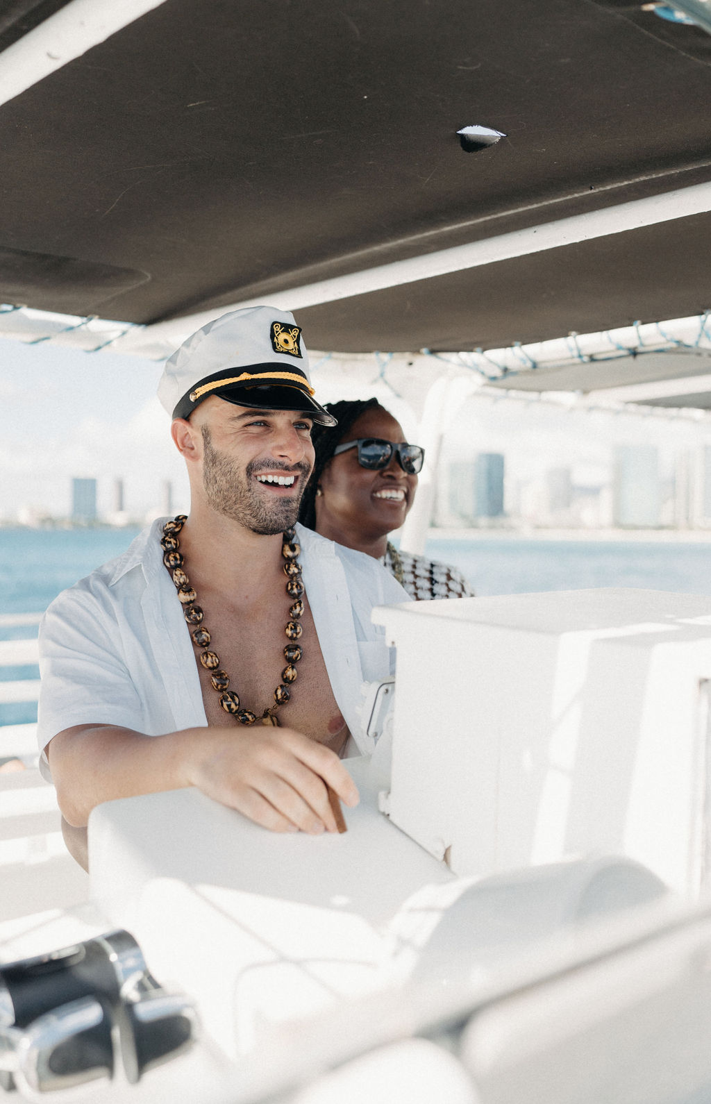 Two people smiling on a boat. One wears a captain's hat and a necklace, steering the vessel. Water and a city's skyline are visible in the background for a micro wedding in oahu