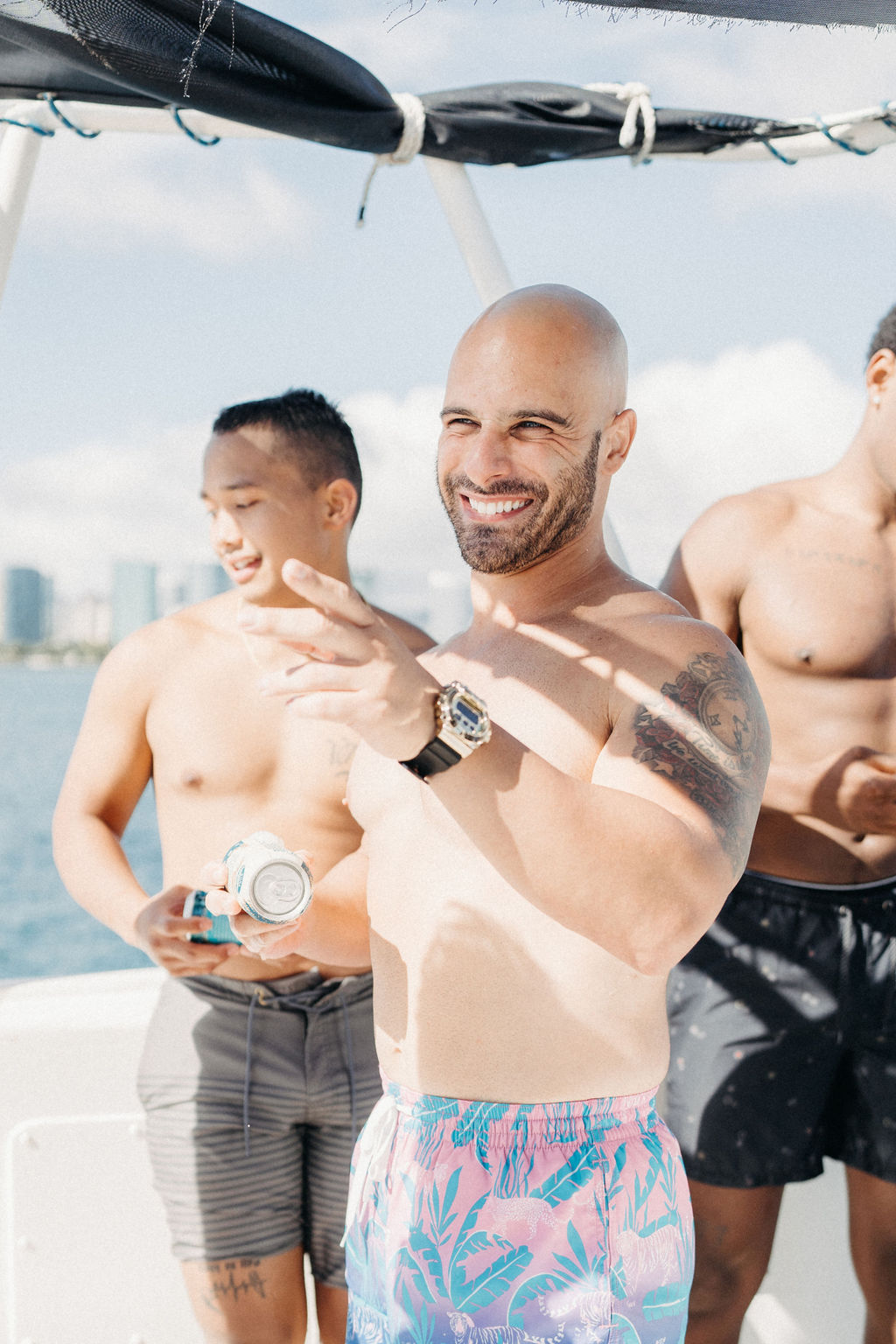 Three men stand on a boat in swim shorts, smiling and enjoying the sunny weather. One man gestures with his hand. The sea and a city skyline are visible in the background for a Micro Wedding in Oahu 