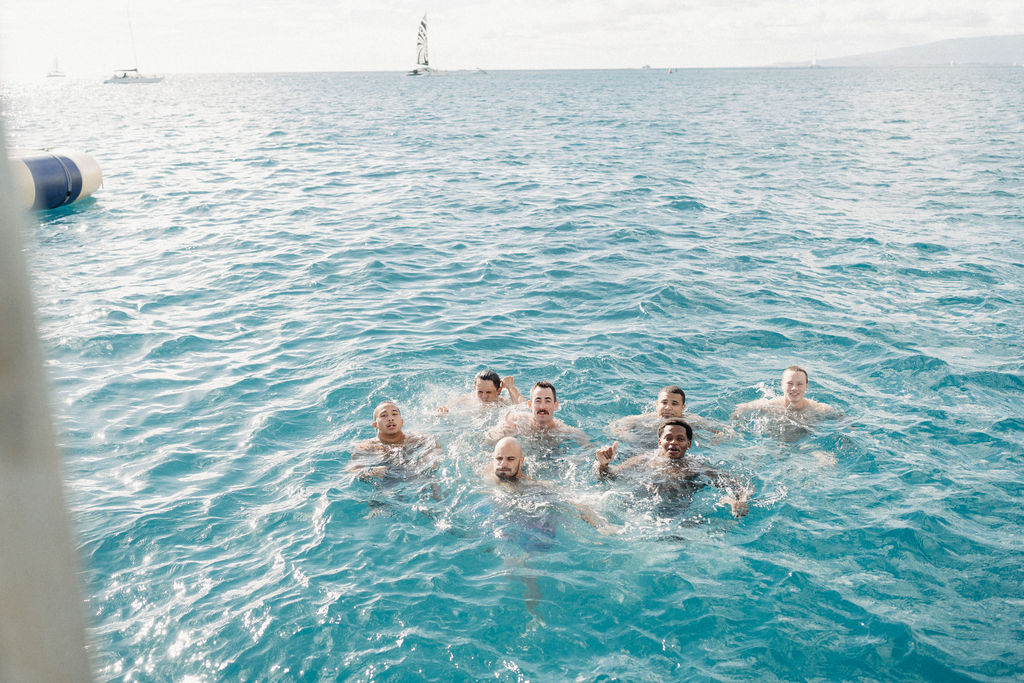 Seven people swimming in the ocean, with boats visible in the distance under a clear sky.