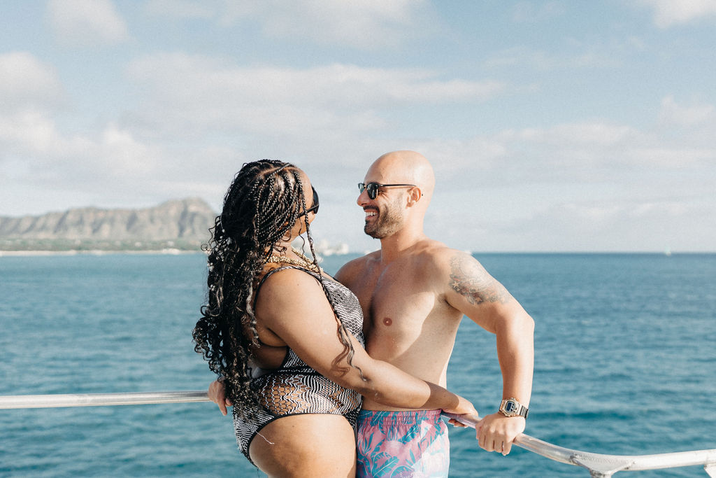 A couple, wearing swimwear, stands on a boat against an ocean backdrop. The man is bald with sunglasses; the woman has braided hair. Mountains are visible in the distance under a partly cloudy sky for a  Micro Wedding in Oahu 