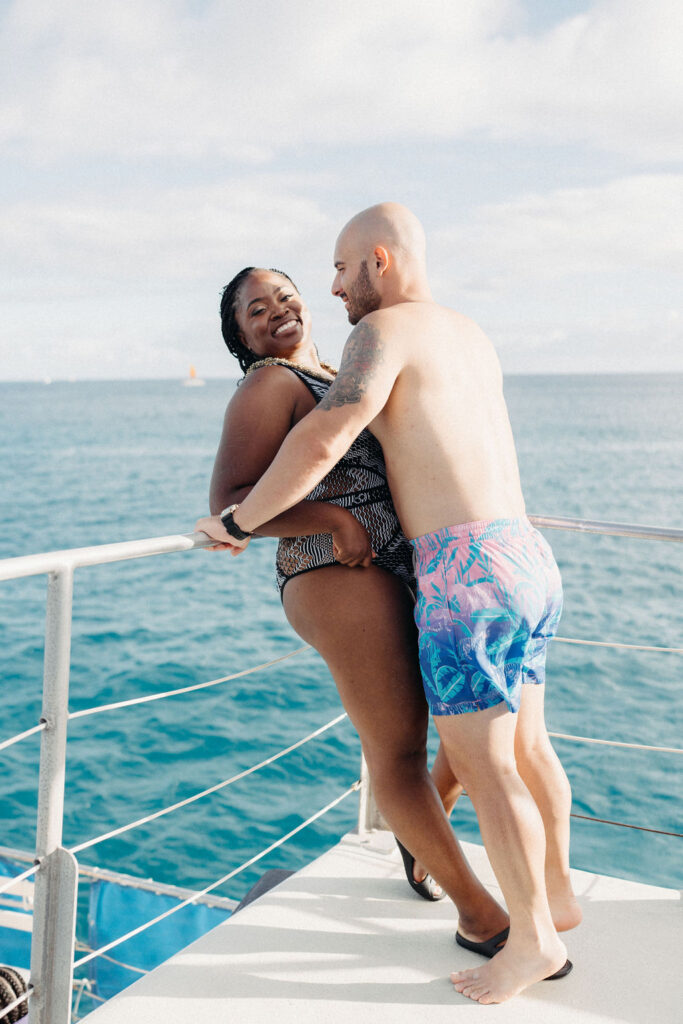 A couple, wearing swimwear, stands on a boat against an ocean backdrop. The man is bald with sunglasses; the woman has braided hair. Mountains are visible in the distance under a partly cloudy sky for a  Micro Wedding in Oahu 