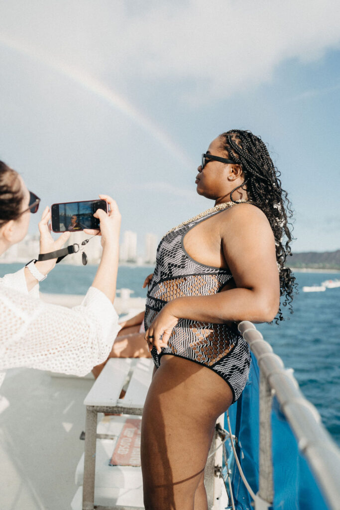 Person in a patterned swimsuit stands on a boat deck, gazing at the ocean. Another person takes their photo with a smartphone. A faint rainbow is visible in the sky.