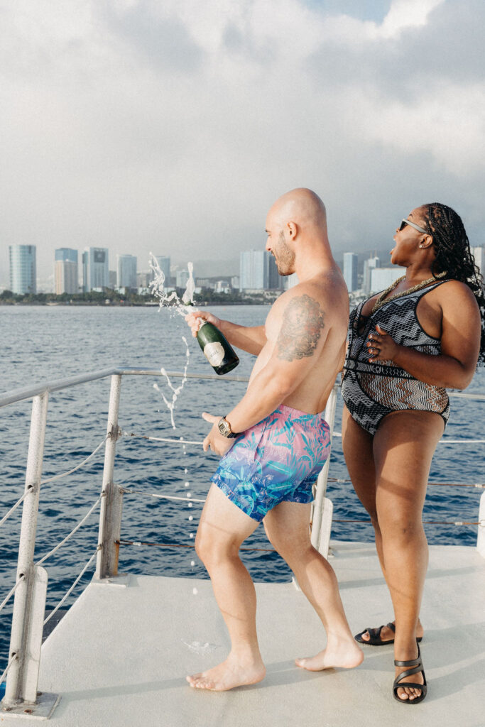 A man in patterned shorts sprays champagne on a boat while a woman in a swimsuit stands nearby. City skyline is visible in the background over the water.