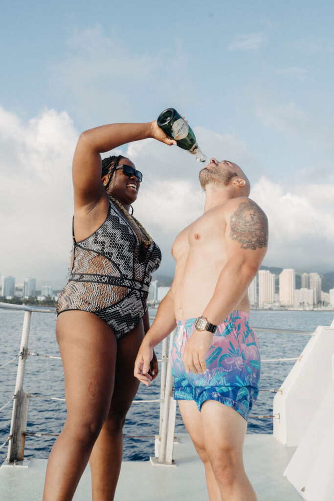 A person in a swimsuit pours champagne into the mouth of another person in swim trunks on a boat, with a city skyline in the background.