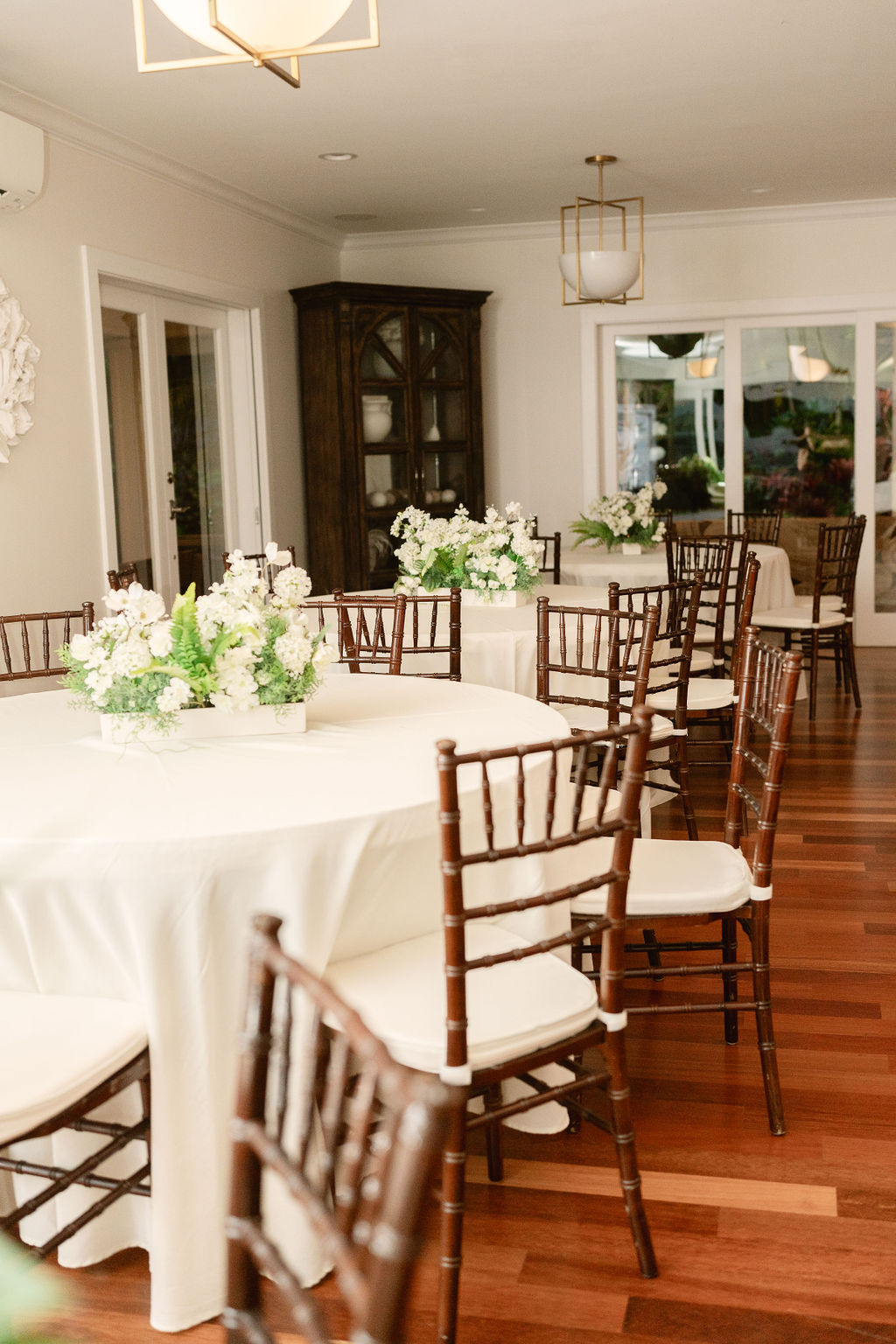 Room with round tables covered in white cloths, surrounded by wooden chairs. Floral centerpieces adorn each table. Hardwood floors and a cabinet are visible in the background.