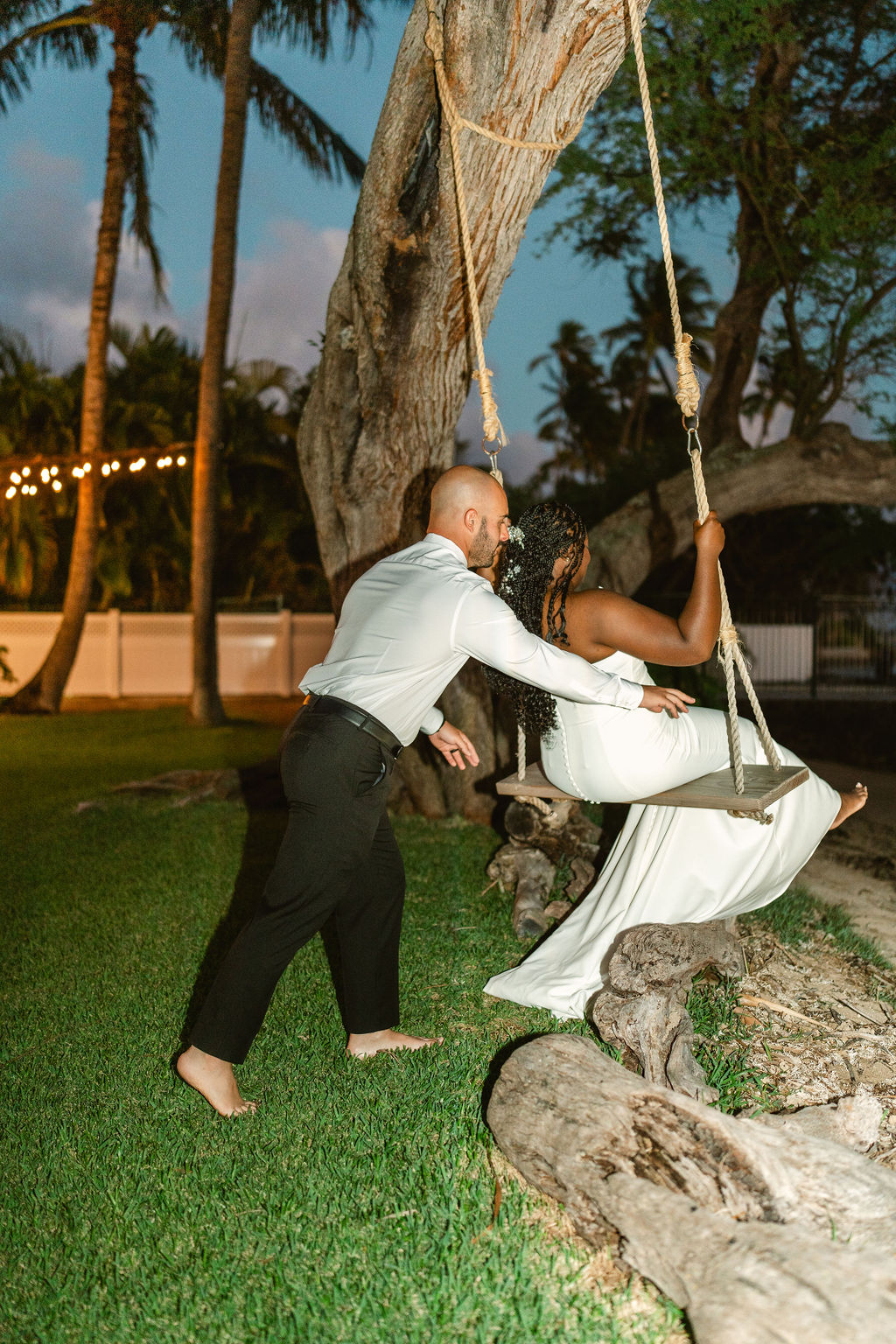 A couple sits on a swing under a large tree, dressed in a wedding gown and suit, holding hands and smiling on a beach.