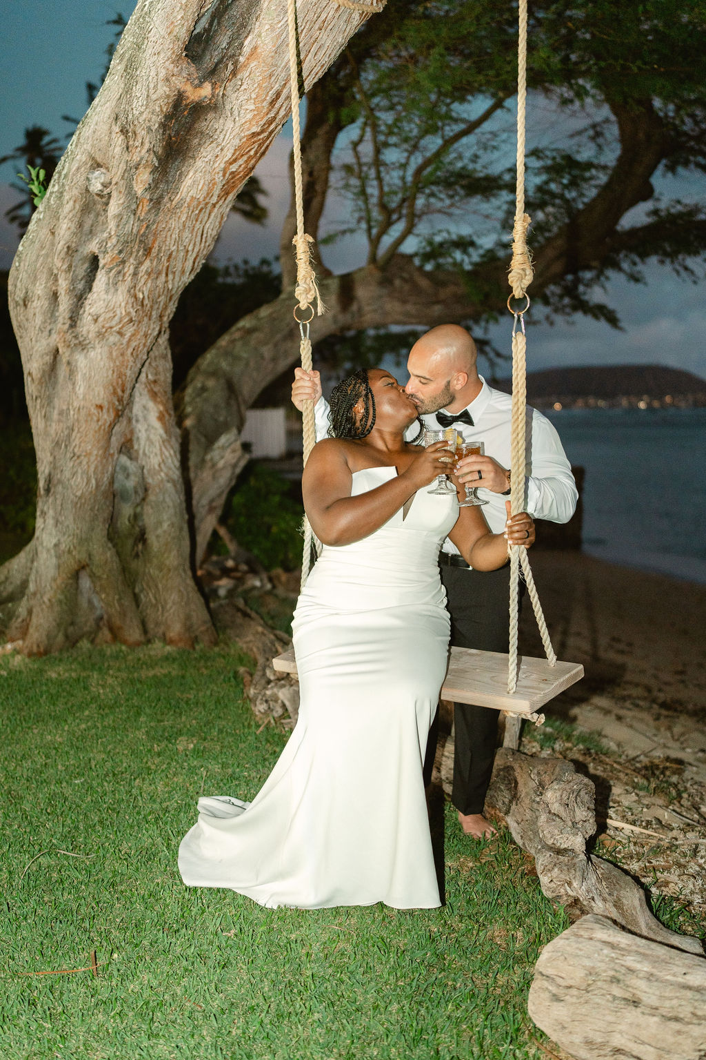 A couple sits on a swing under a large tree, dressed in a wedding gown and suit, holding hands and smiling on a beach.